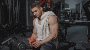 Sportininkas sporto salėje sėdi ir laiko proteino gertuvę.
